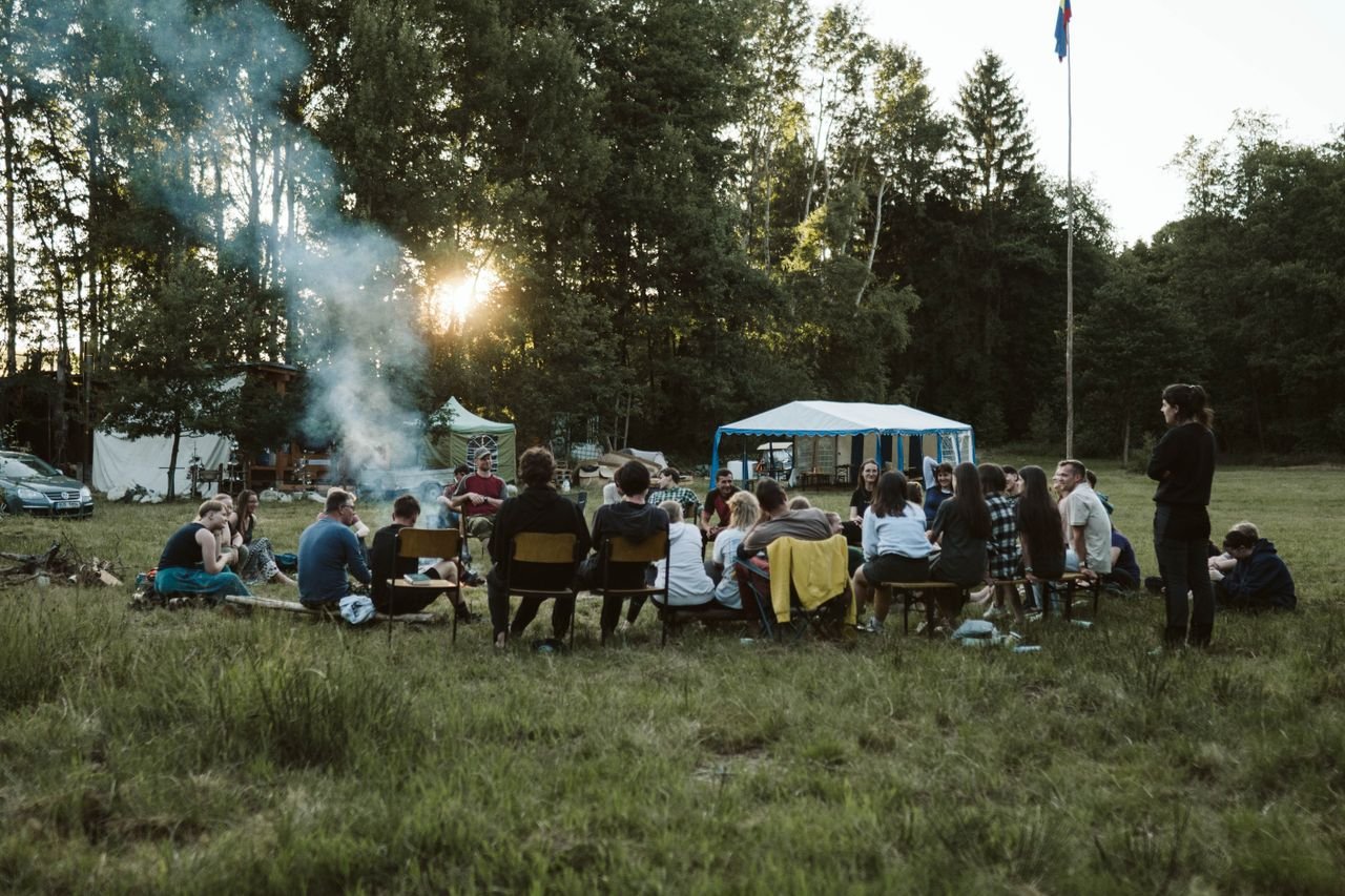 Home A group of adults relaxing around a campfire in a scenic outdoor setting with tents.
